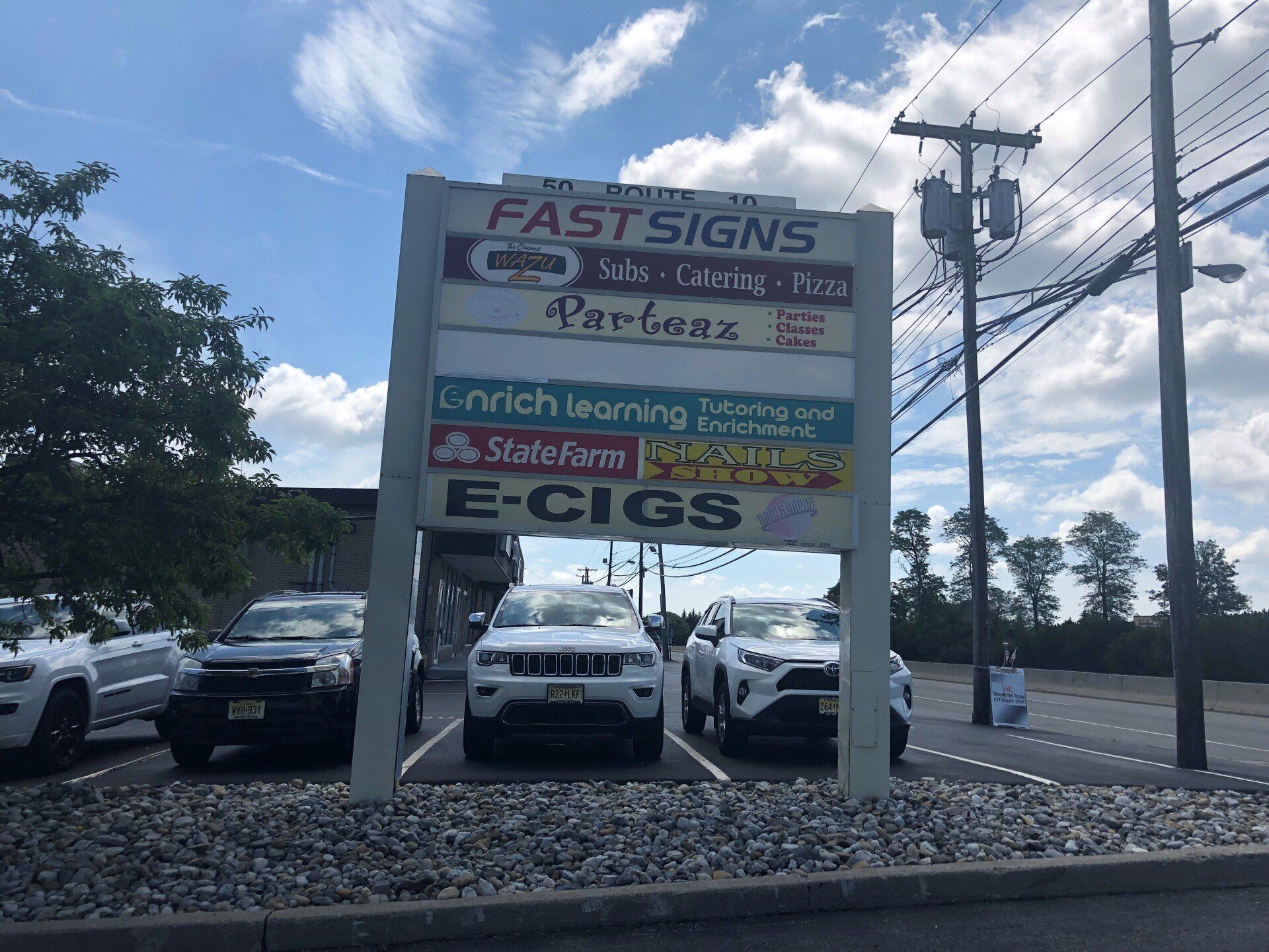 Sign for a strip mall, with businesses including FastSigns, Parlour, and E-CIGS. Cars parked out front.