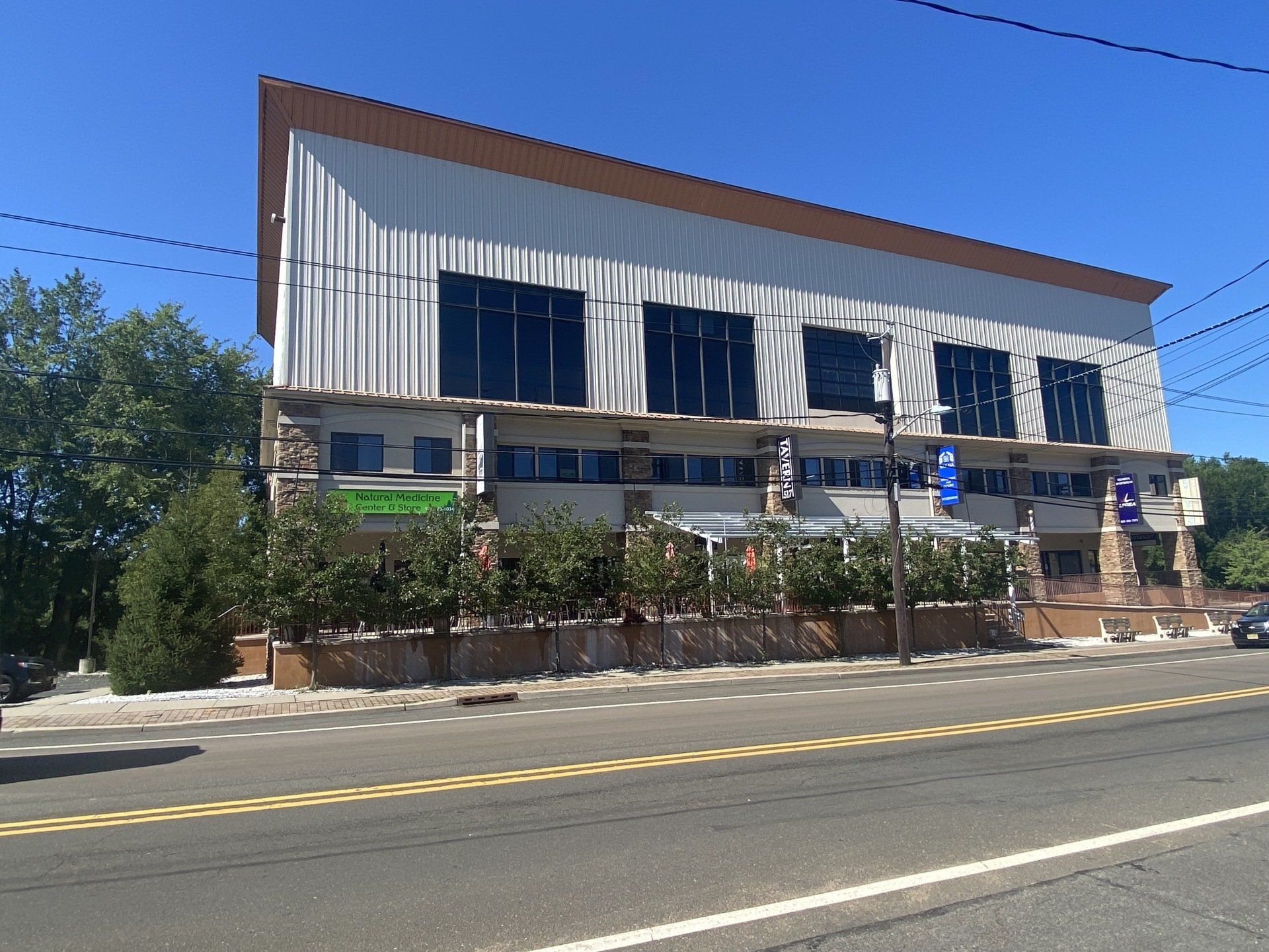 Two-story commercial building with light and dark facades, large windows, and a brown roof. Road in the foreground, blue sky.