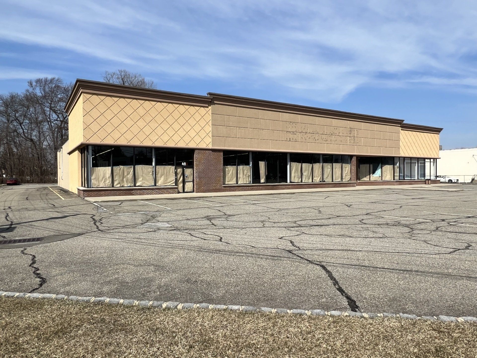Vacant, single-story commercial building with large windows and a cracked parking lot. Beige facade, under a blue sky.
