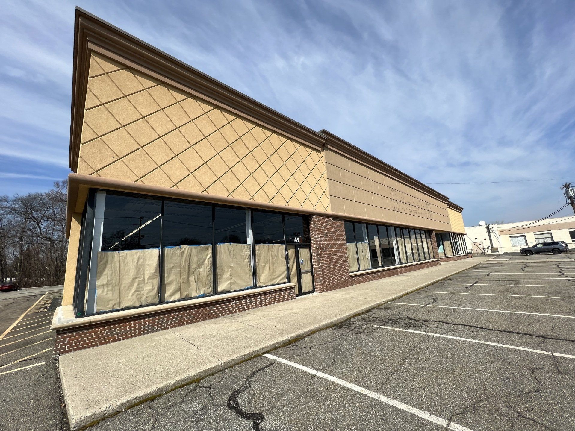 Empty commercial building with large windows and tan facade, in an asphalt parking lot under a cloudy sky.