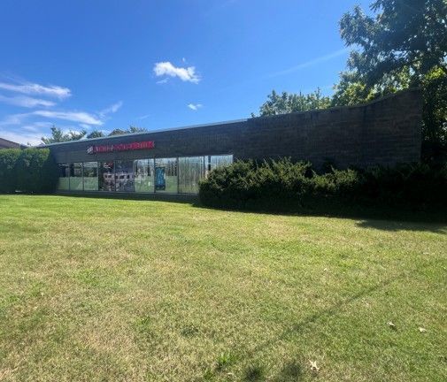 Single-story storefront with red signage, glass windows, and a grassy lawn under a bright blue sky.