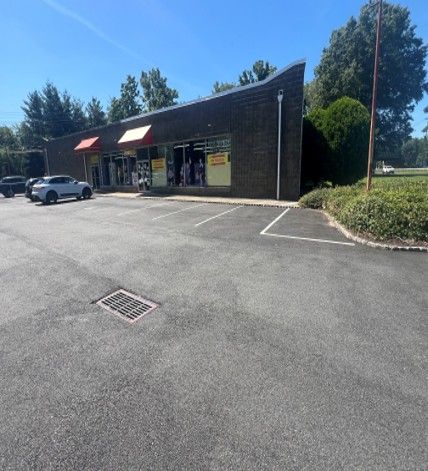 Sunlit parking lot beside a building, with cars parked along a curved asphalt drive and trees in the background
