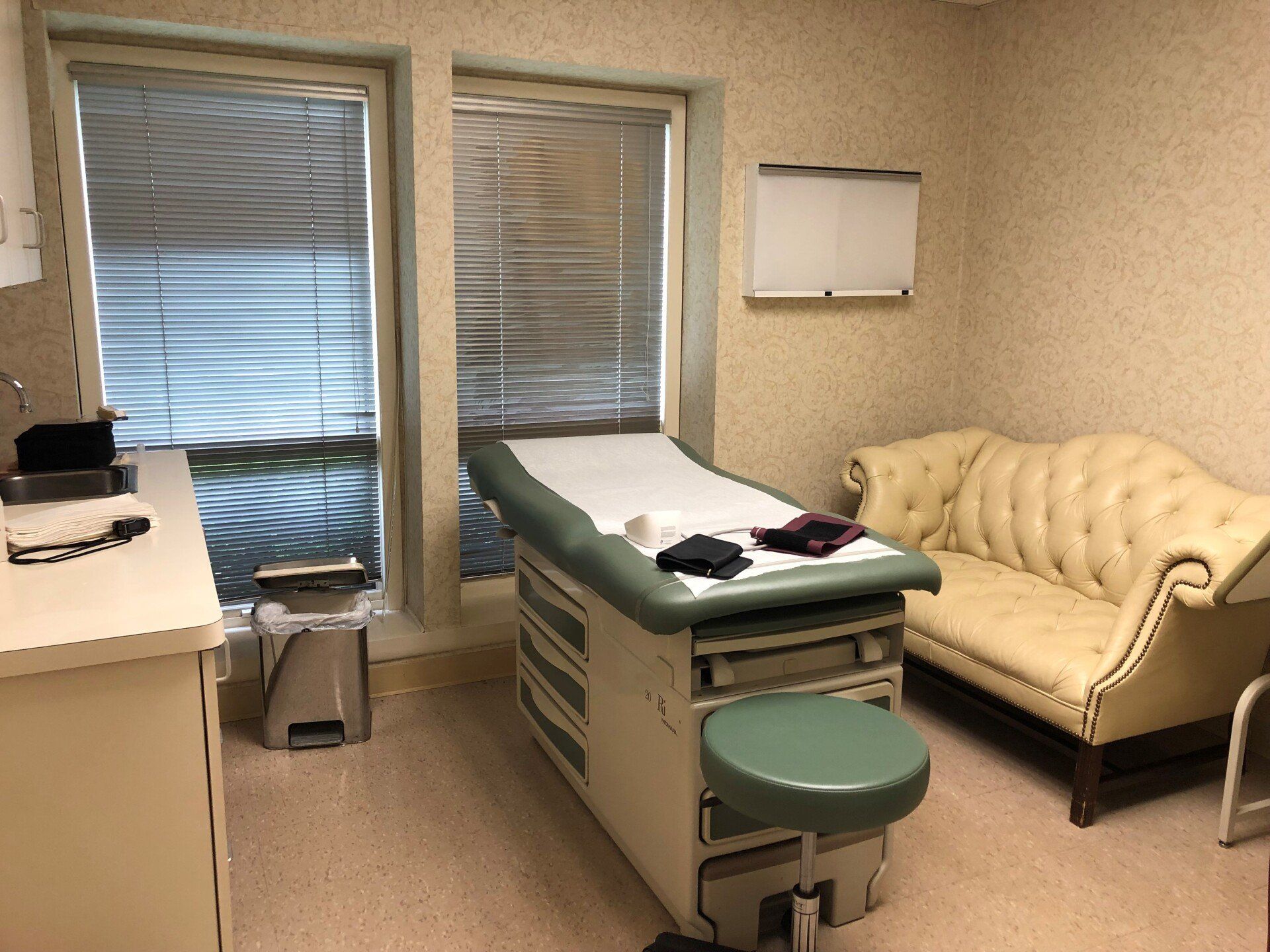 Doctor's examination room with exam table, couch, windows with blinds, and medical equipment.