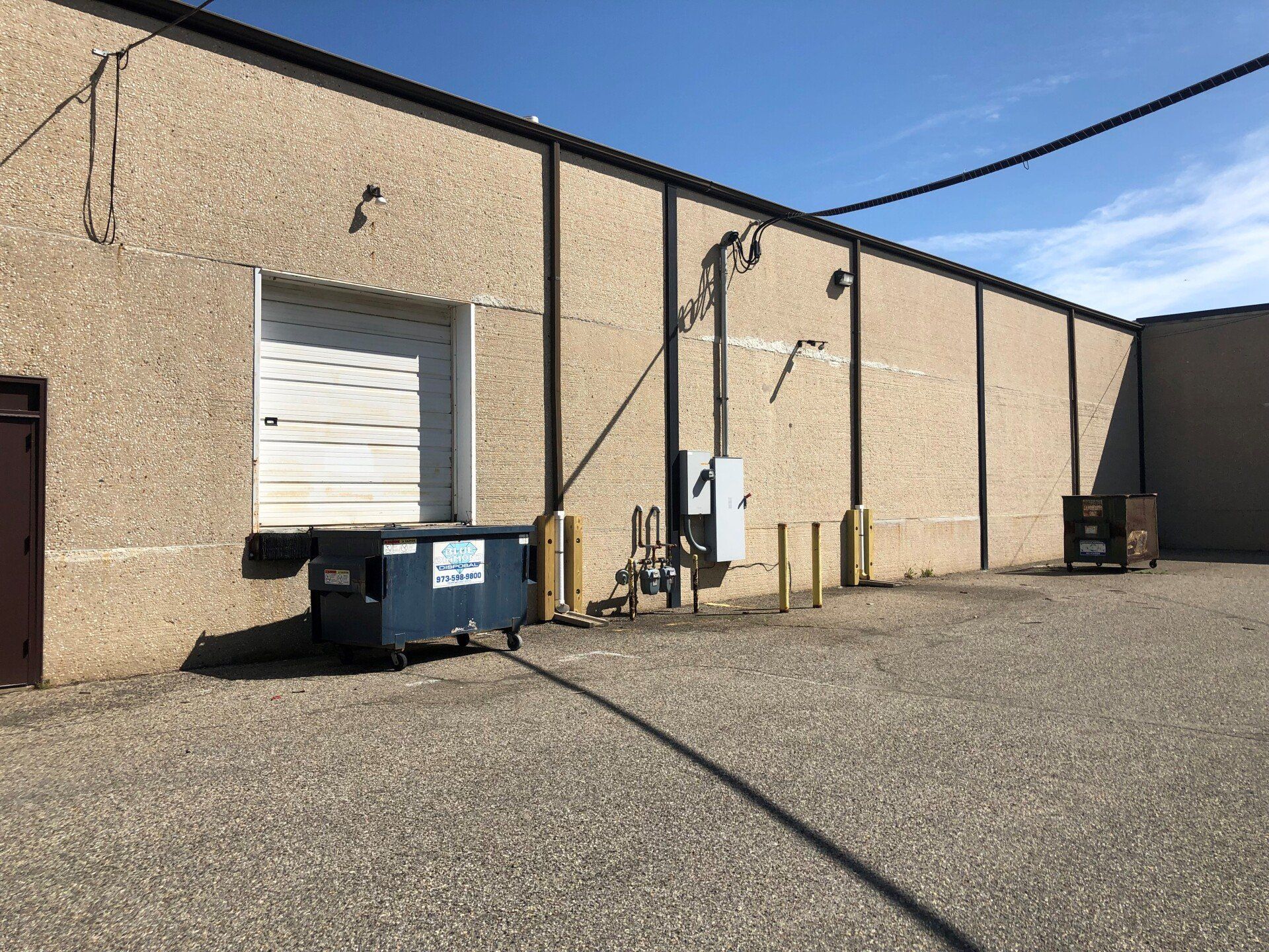 Exterior view of a commercial building with a loading dock, dumpster, and utility box on a gravel lot.