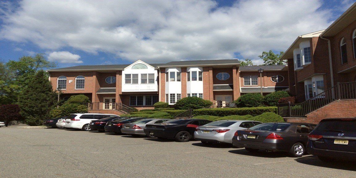 A multi-story brick commercial building with white trim sits behind a gravel parking lot filled with parked cars.