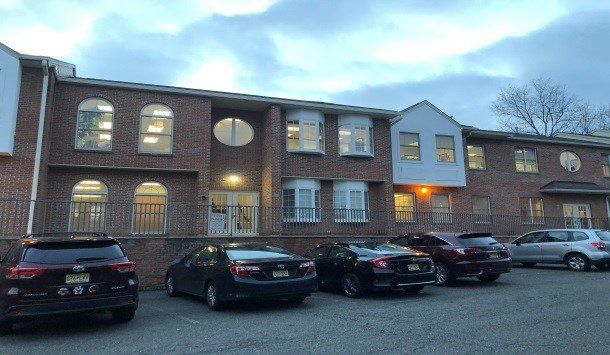 Brick building with multiple windows. Cars parked in front. Cloudy sky.
