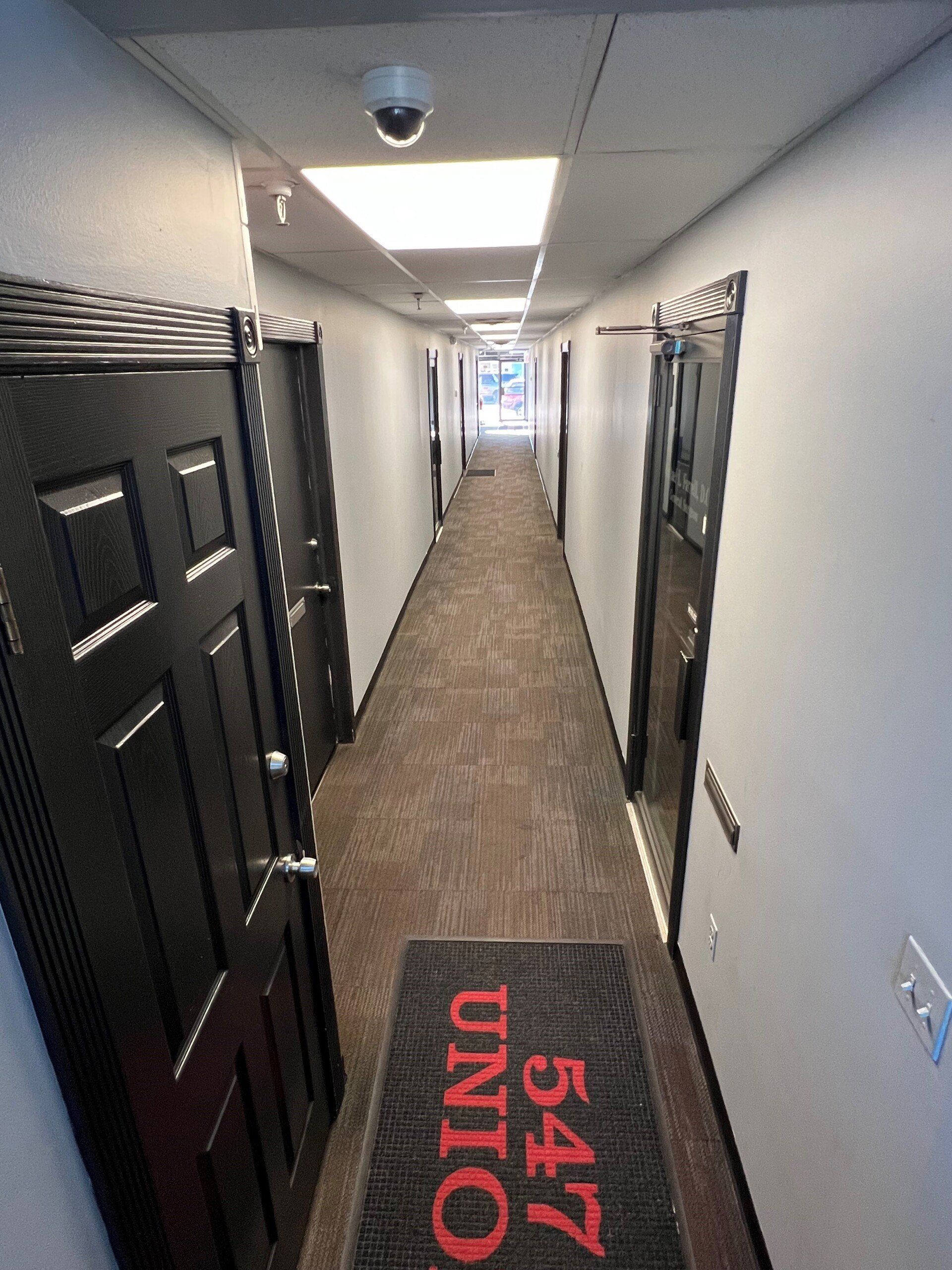 Narrow hallway with dark doors, brown carpet, overhead lighting, security camera. Red welcome mat with “547 UNION”.