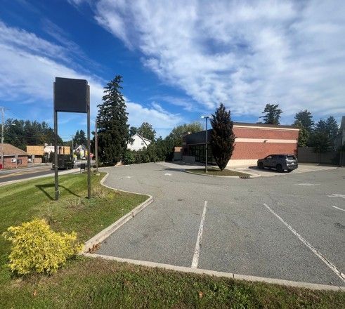 Empty parking lot beside a brick building under a blue sky, with a signpost and trees nearby