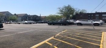 Cars parked in an asphalt lot on a sunny day; buildings and trees in background.