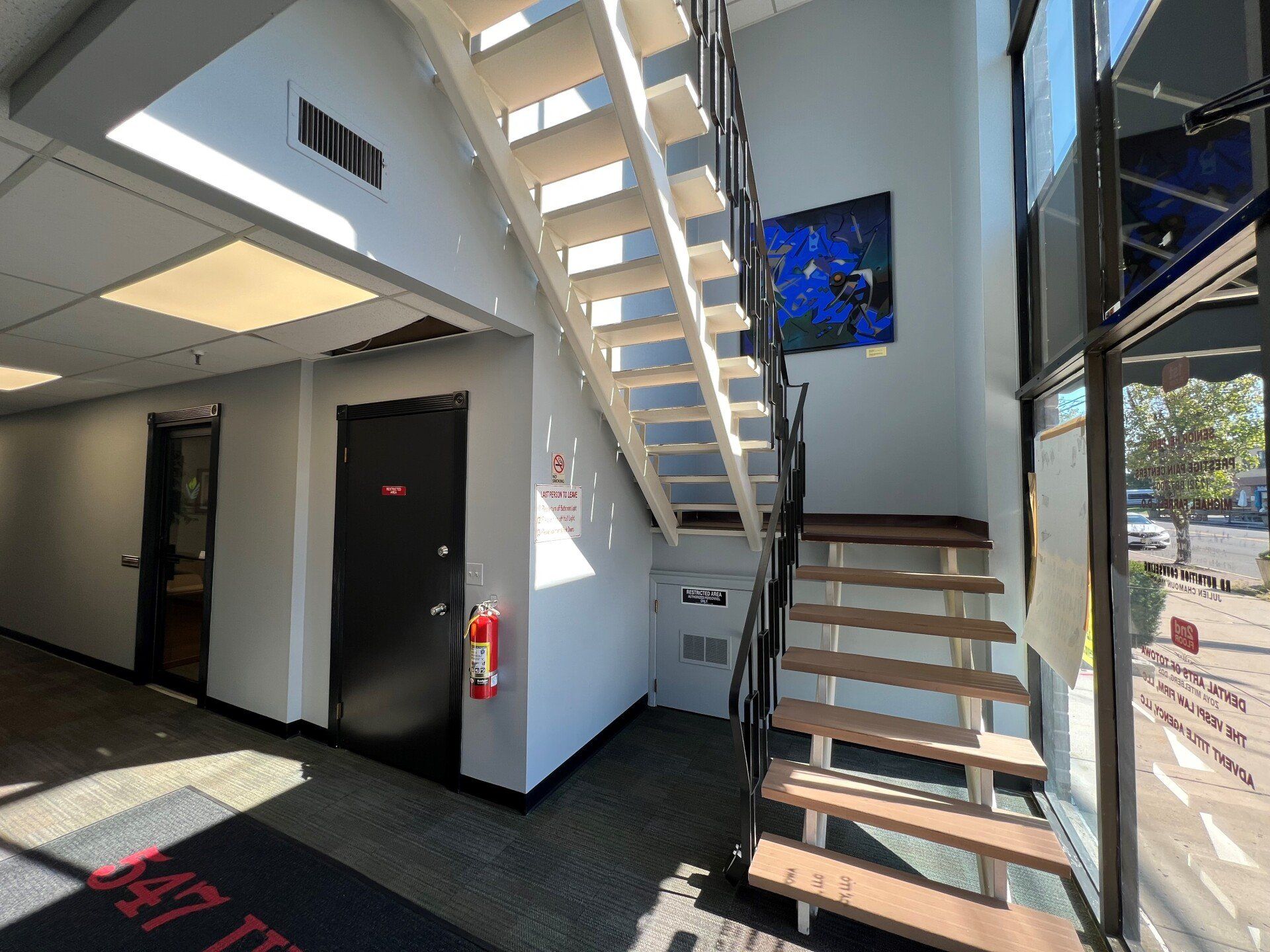 Interior shot with stairs, a door, and a painting on the wall. Sunlight streams in from a window.