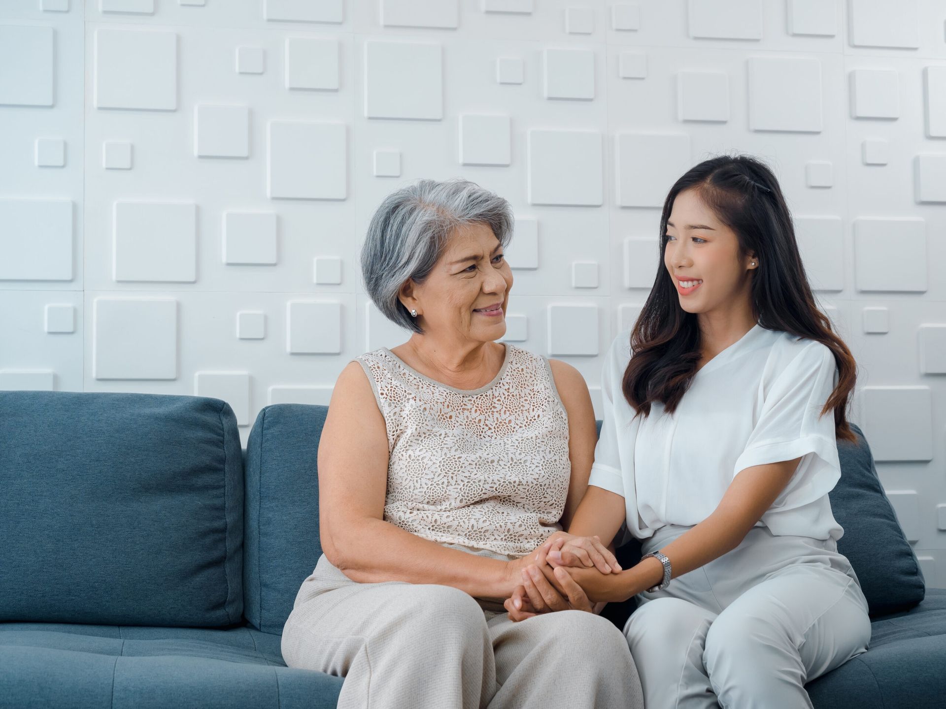 Two women sit on a blue couch, holding hands and smiling in a bright living room.