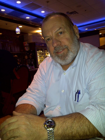 Man seated at a dimly lit bar, smiling slightly, wearing a light blue shirt and a watch.
