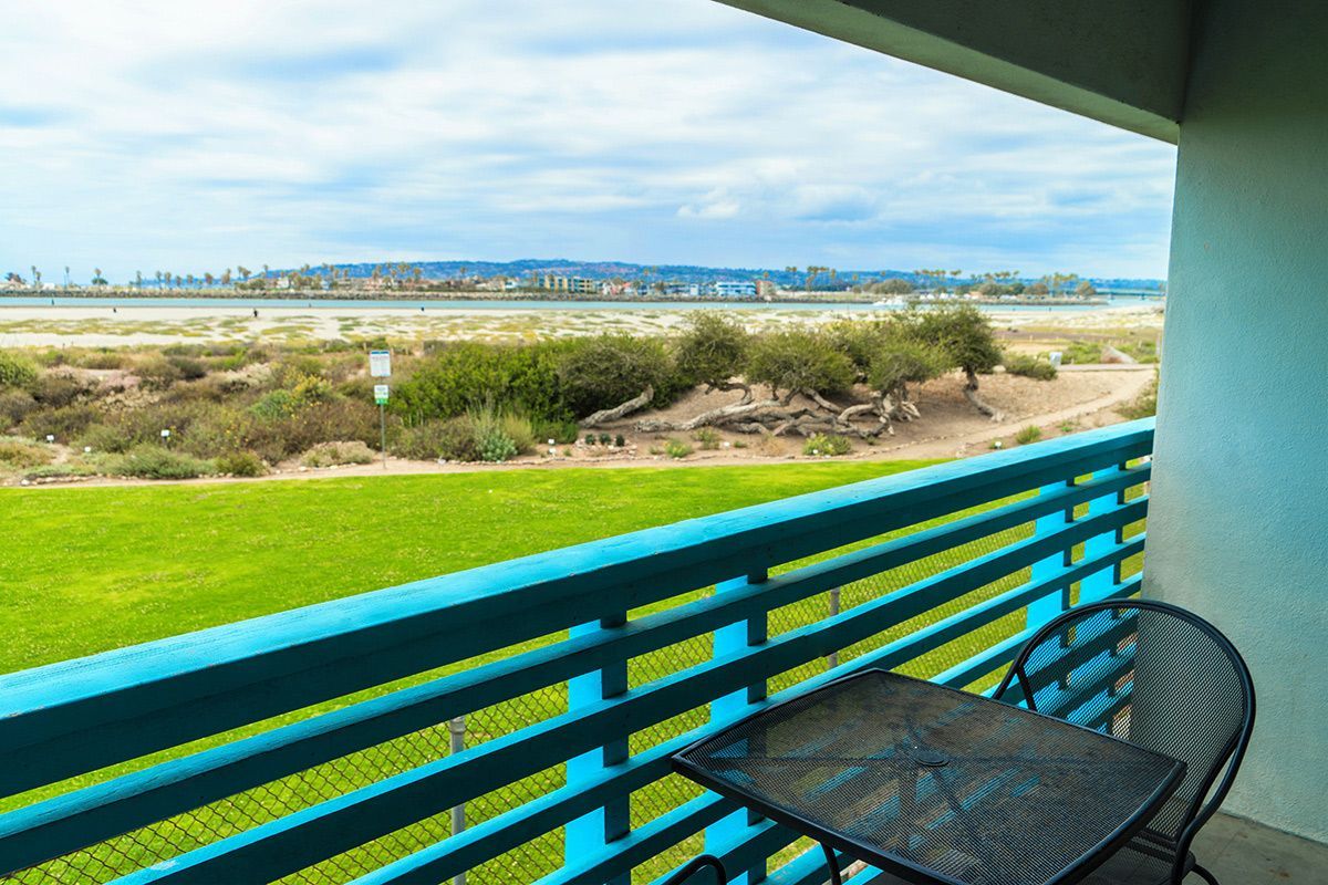 Balcony with turquoise railing, small table and chair, overlooking grassy field and water.