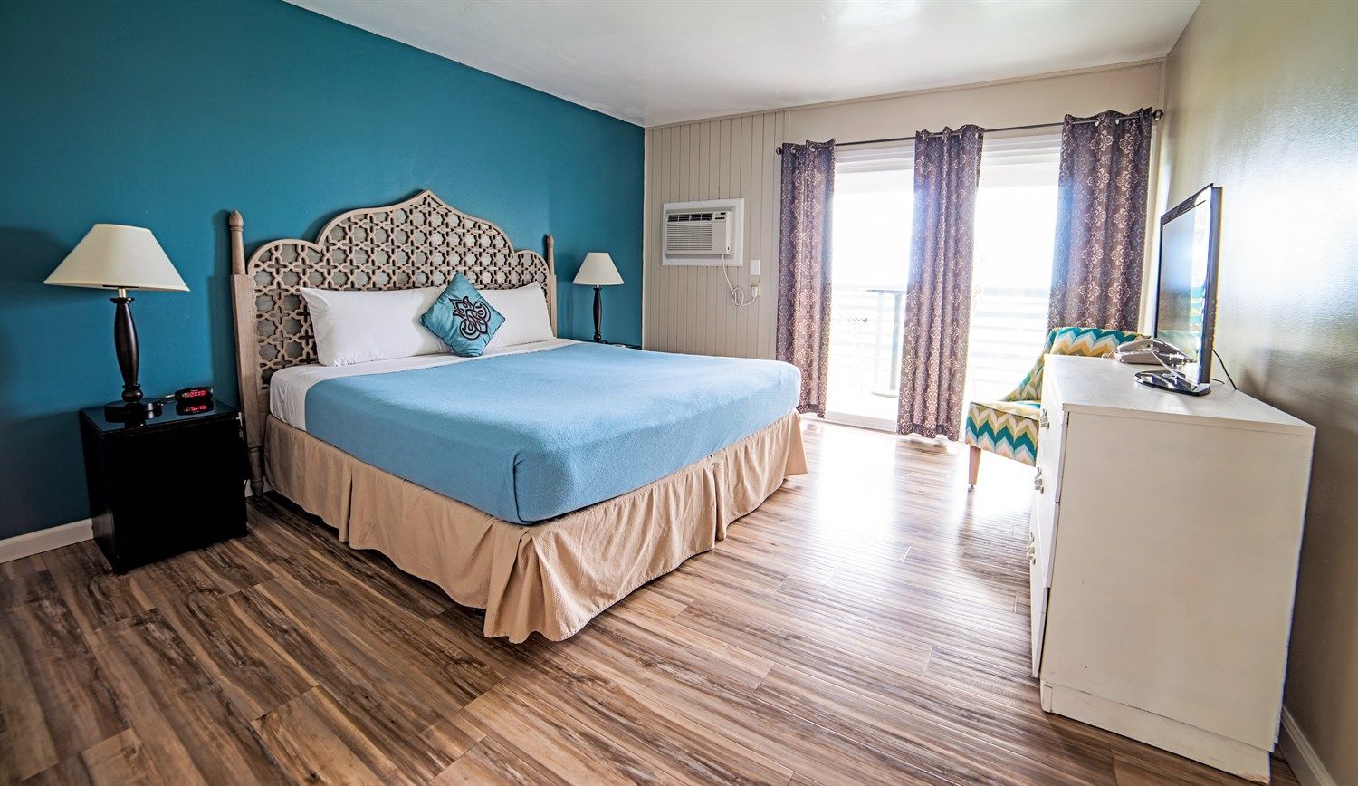 Bedroom with king-sized bed, blue walls, and a balcony. Includes a TV, nightstands, and a patterned chair.