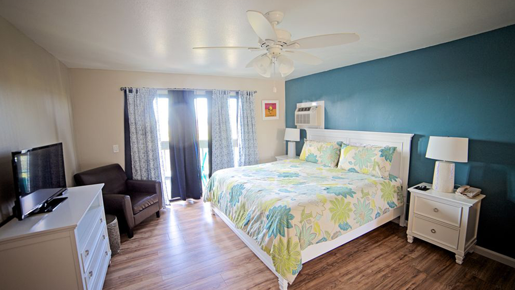 Bedroom with a king-sized bed, a dresser, armchair, and a door to the balcony. Teal accent wall.