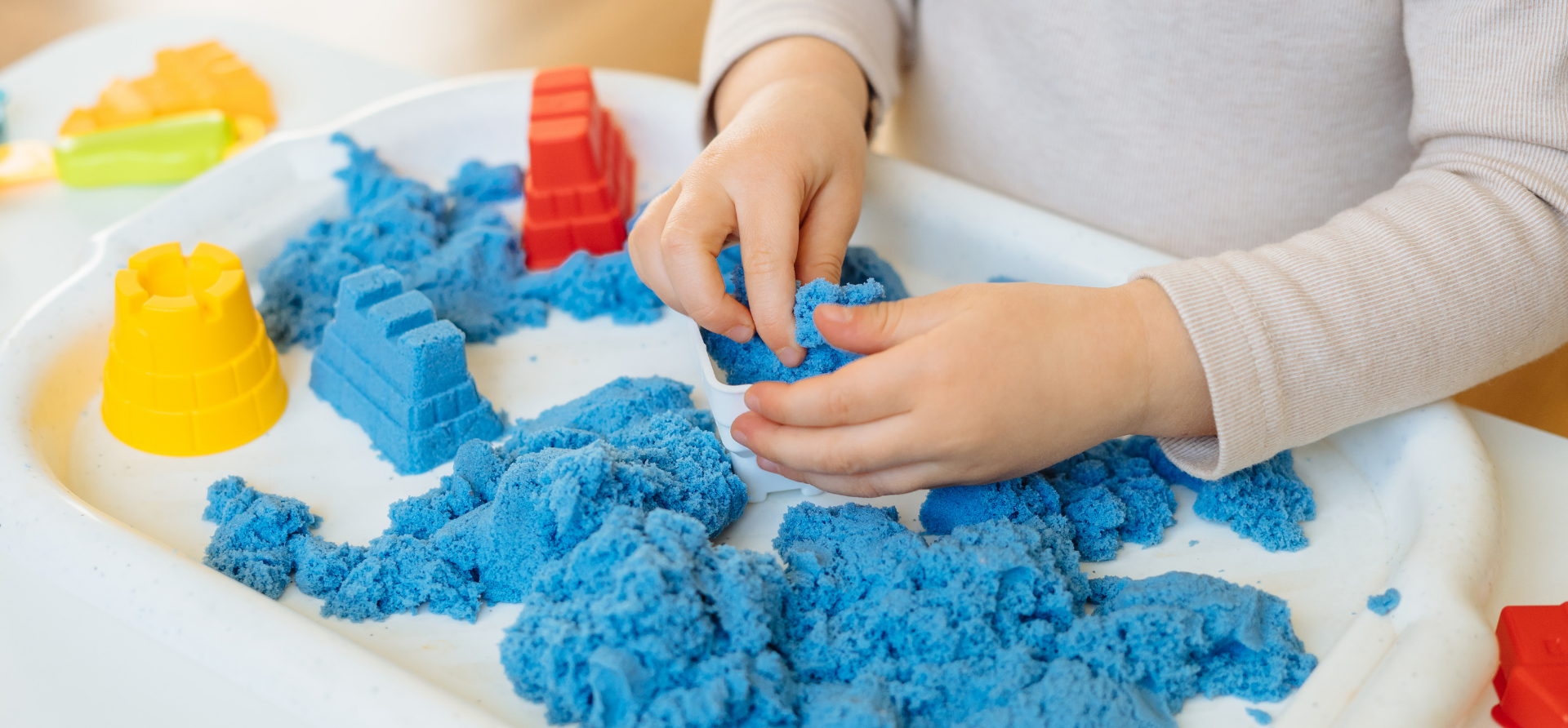 Child playing with blue kinetic sand in a white tray, with yellow and red toy molds.