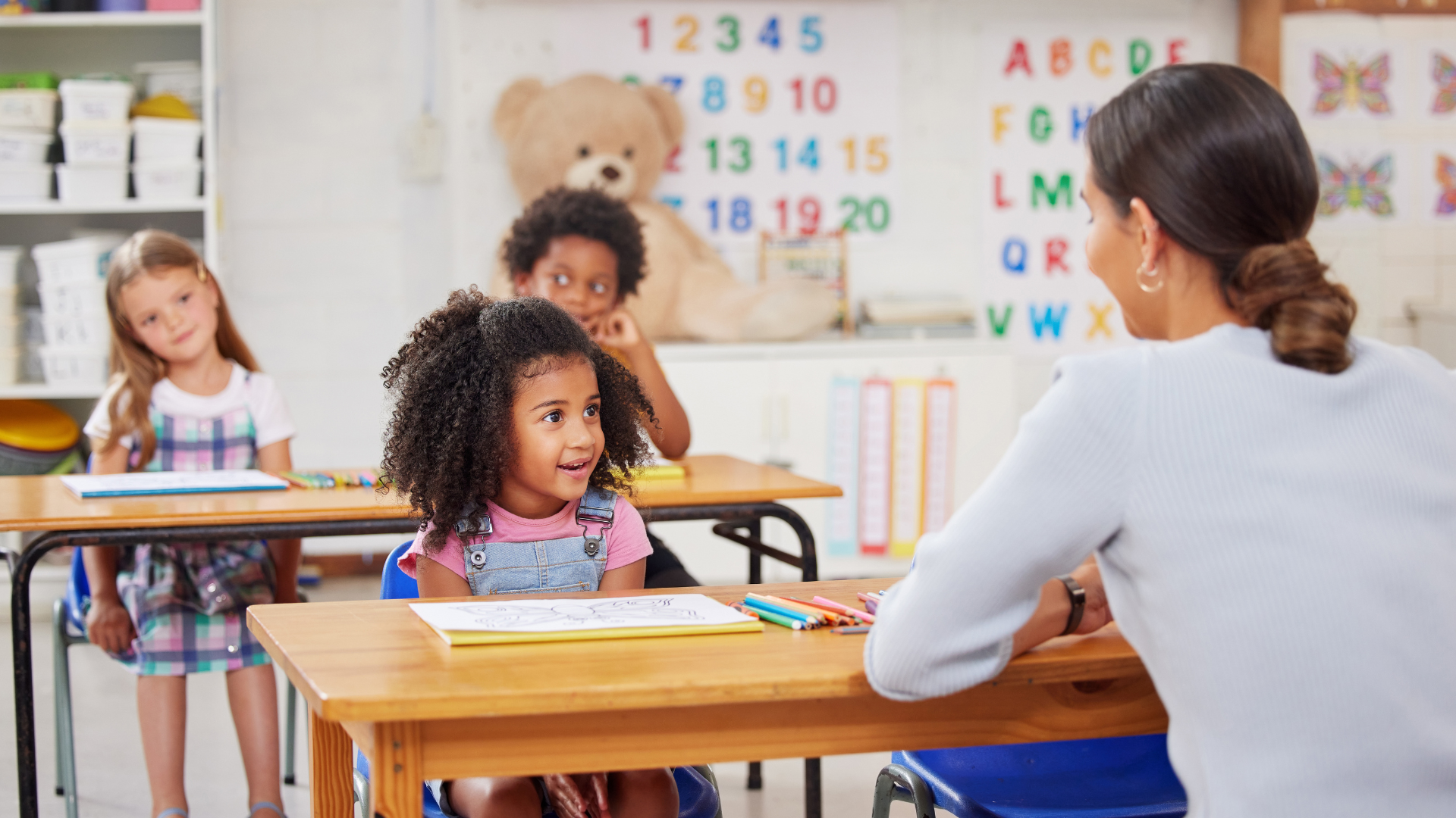 A teacher is sitting at a desk in front of a group of children in a classroom.