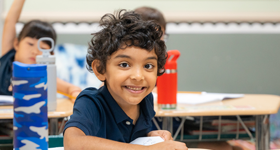 A young boy is smiling while sitting at a desk in a classroom.