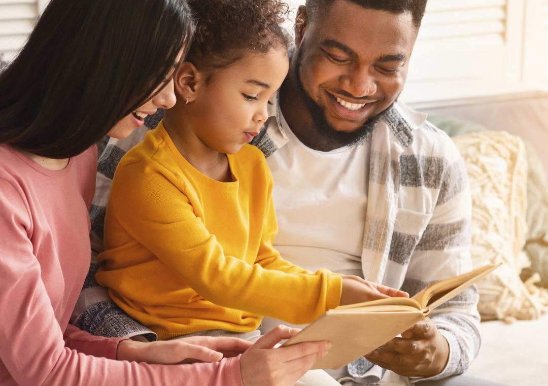Family reading a book together; child in yellow, parents smiling indoors.