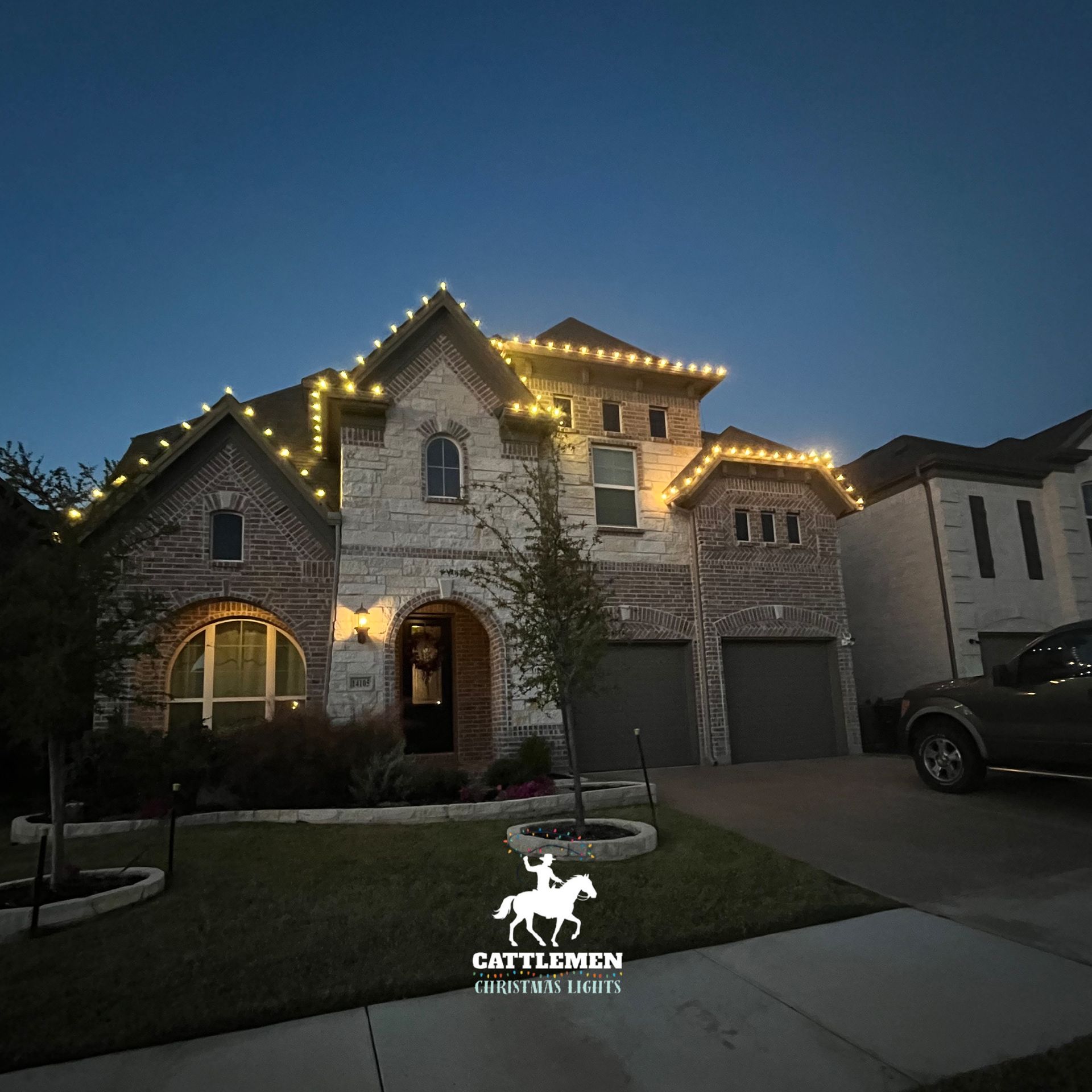 Two-story stone house at dusk with warm roofline lights and a lit front entryway