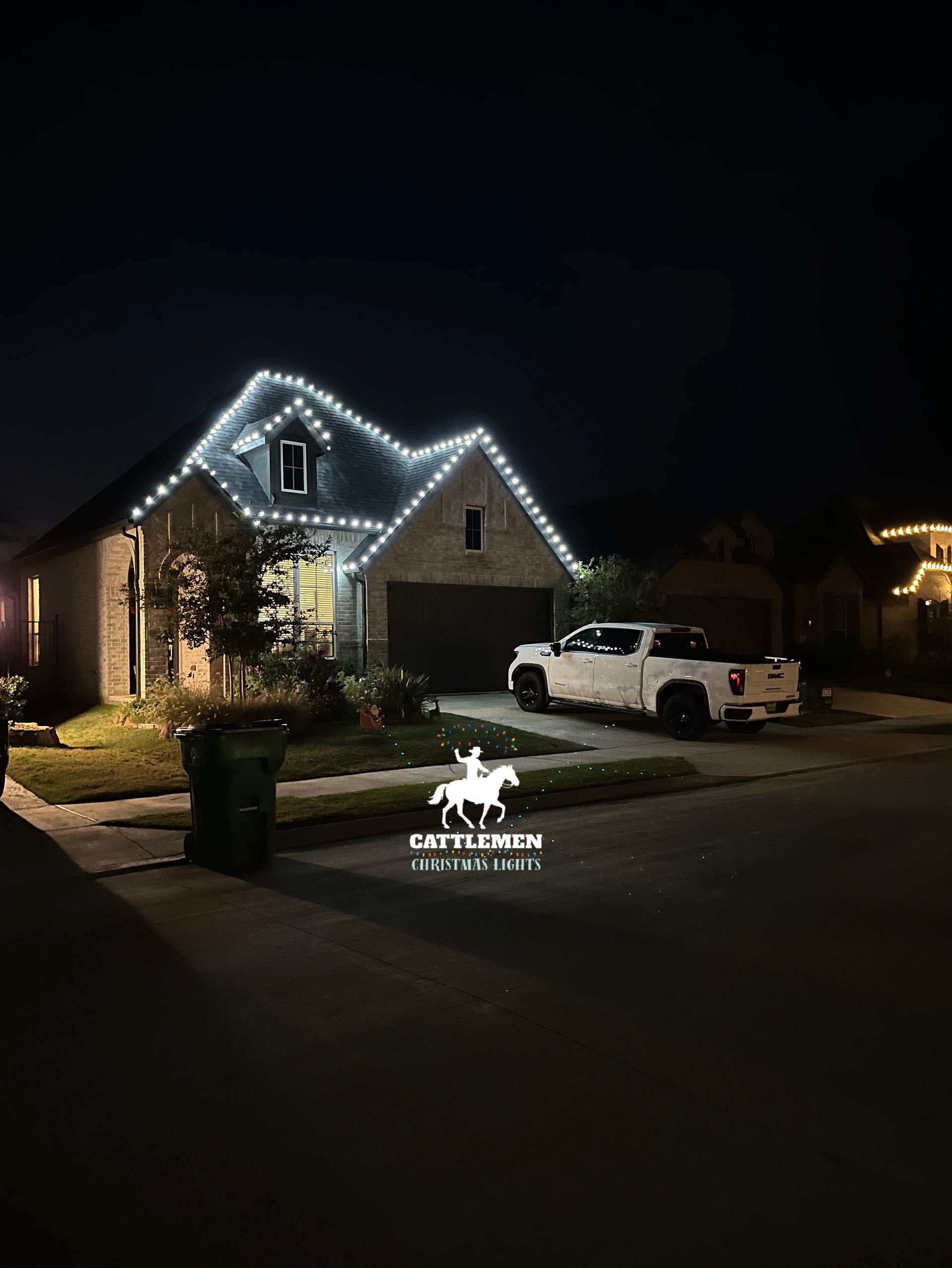 House outlined with white holiday lights at night, with a white pickup truck in the driveway.