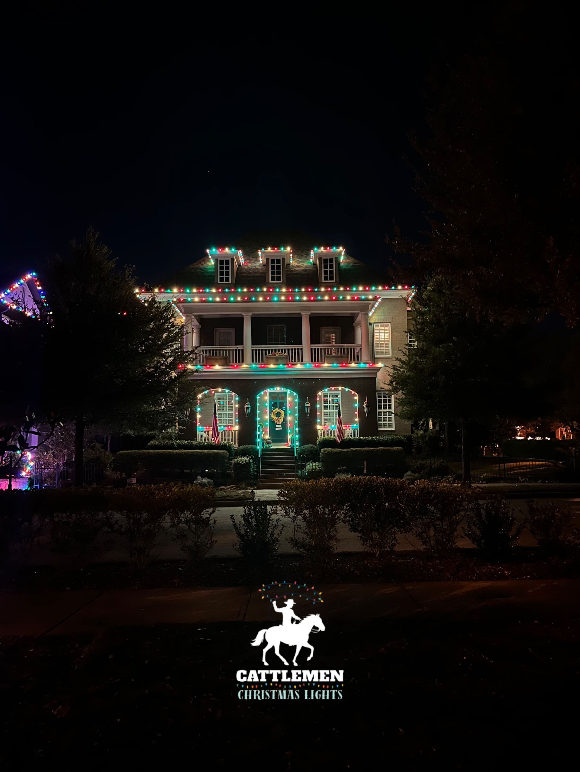 House outlined in colorful holiday lights at night, with a lit sign in front.