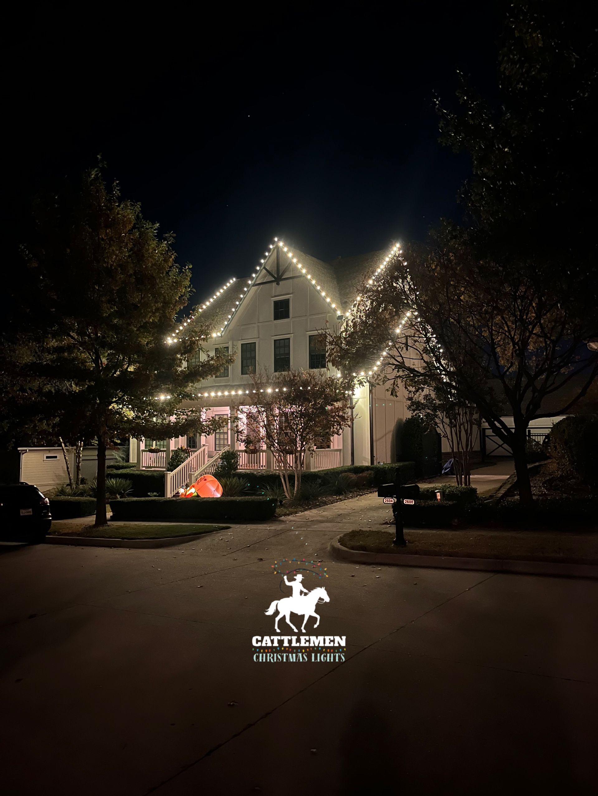 House lit with white holiday lights at night, with illuminated trees and a dark street in front