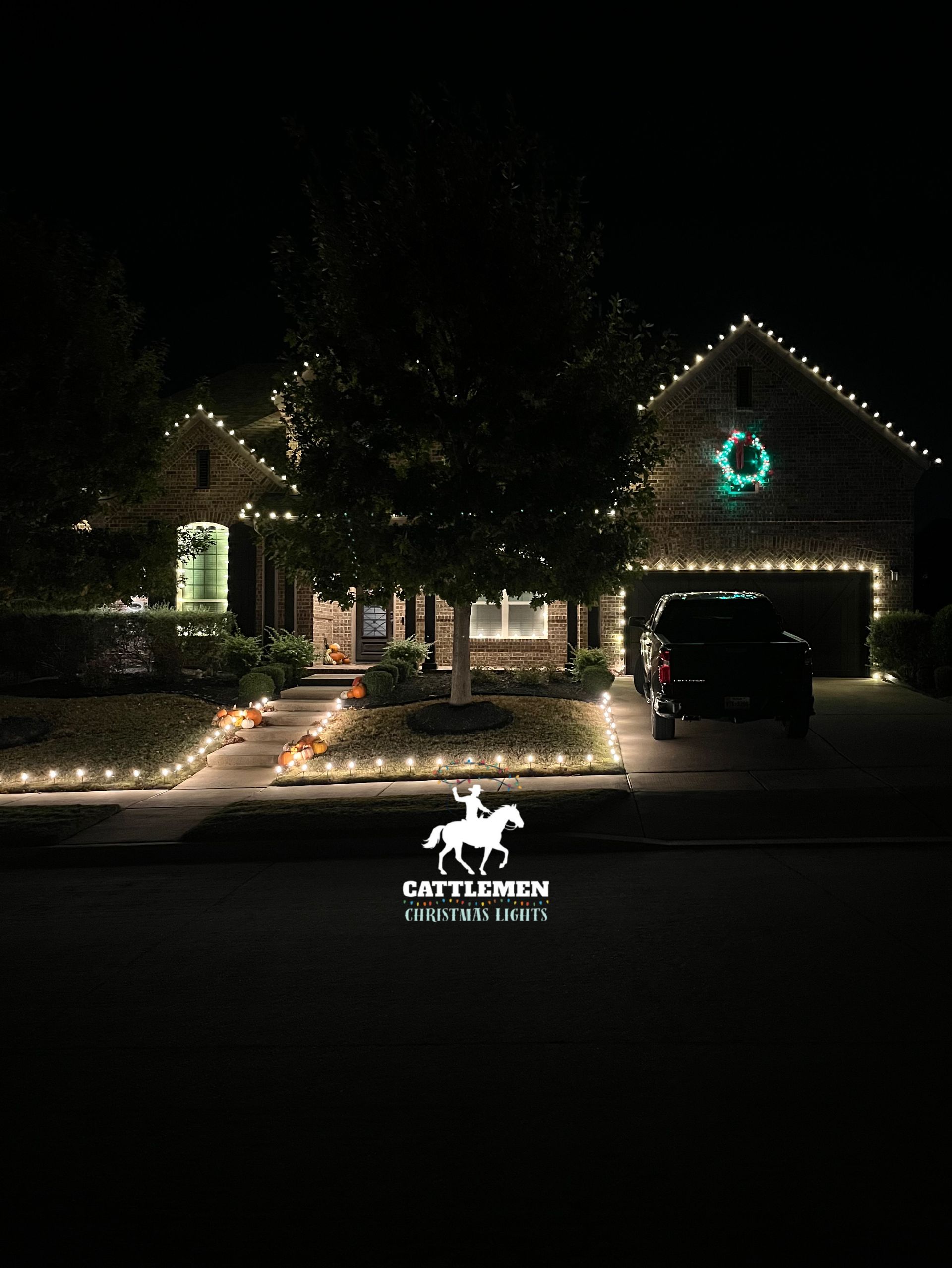House at night decorated with white holiday lights, a lit tree in front, and a parked car in the driveway.