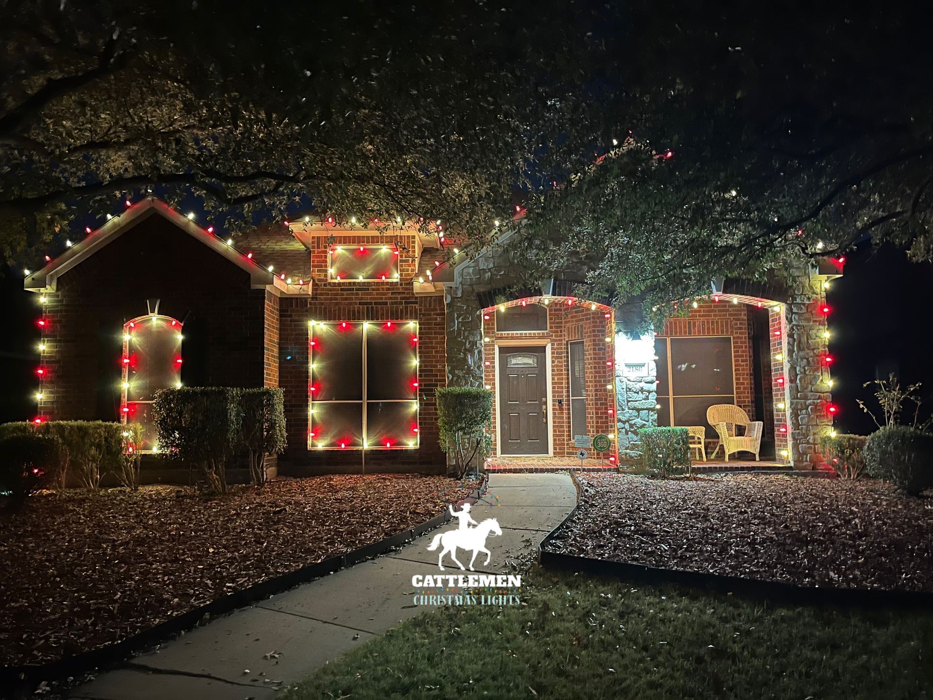 House at night decorated with red and white Christmas lights, with a lit walkway and falling snow.