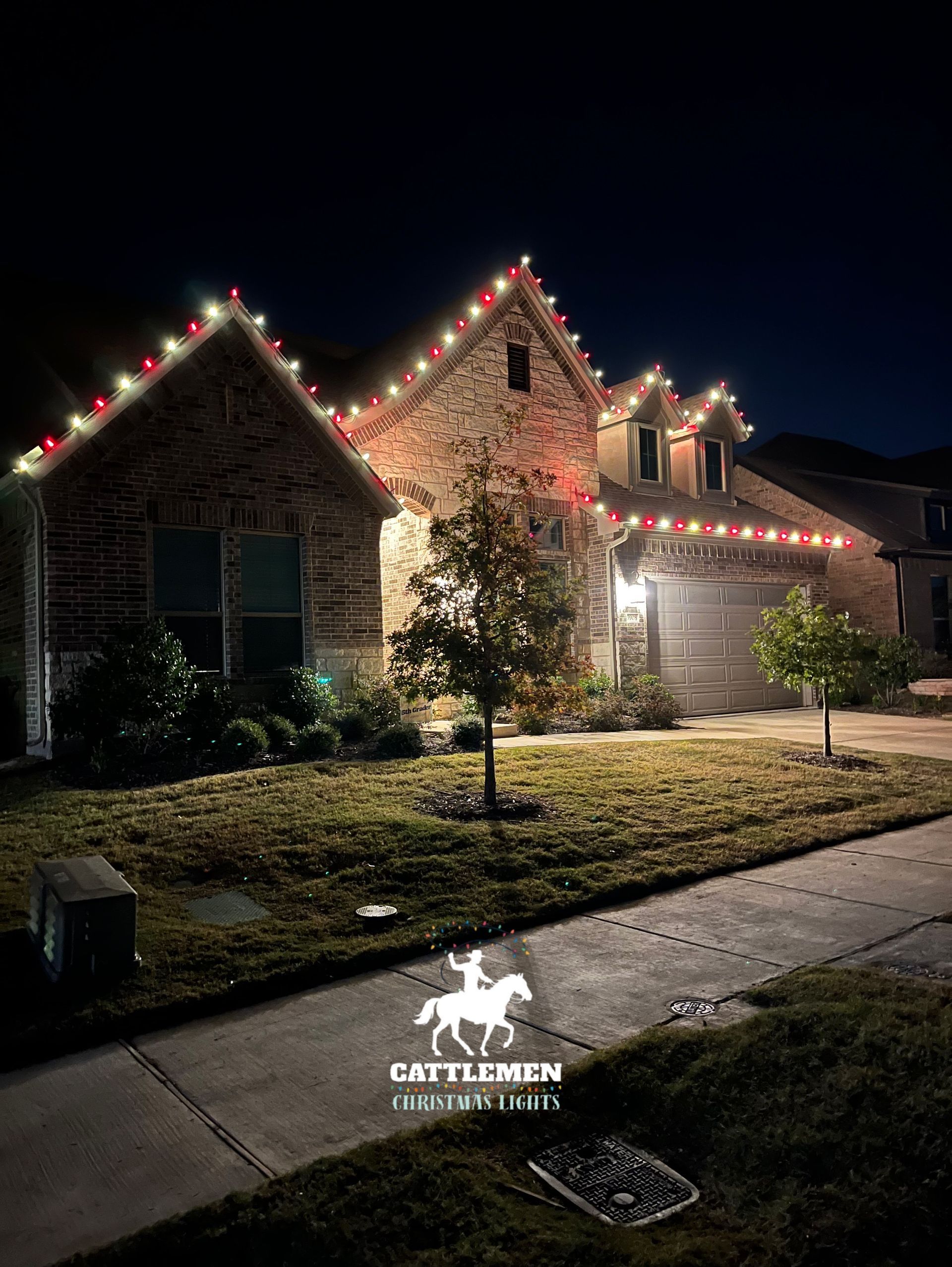 Brick house at night outlined with red and white holiday lights, with a lit driveway and lawn