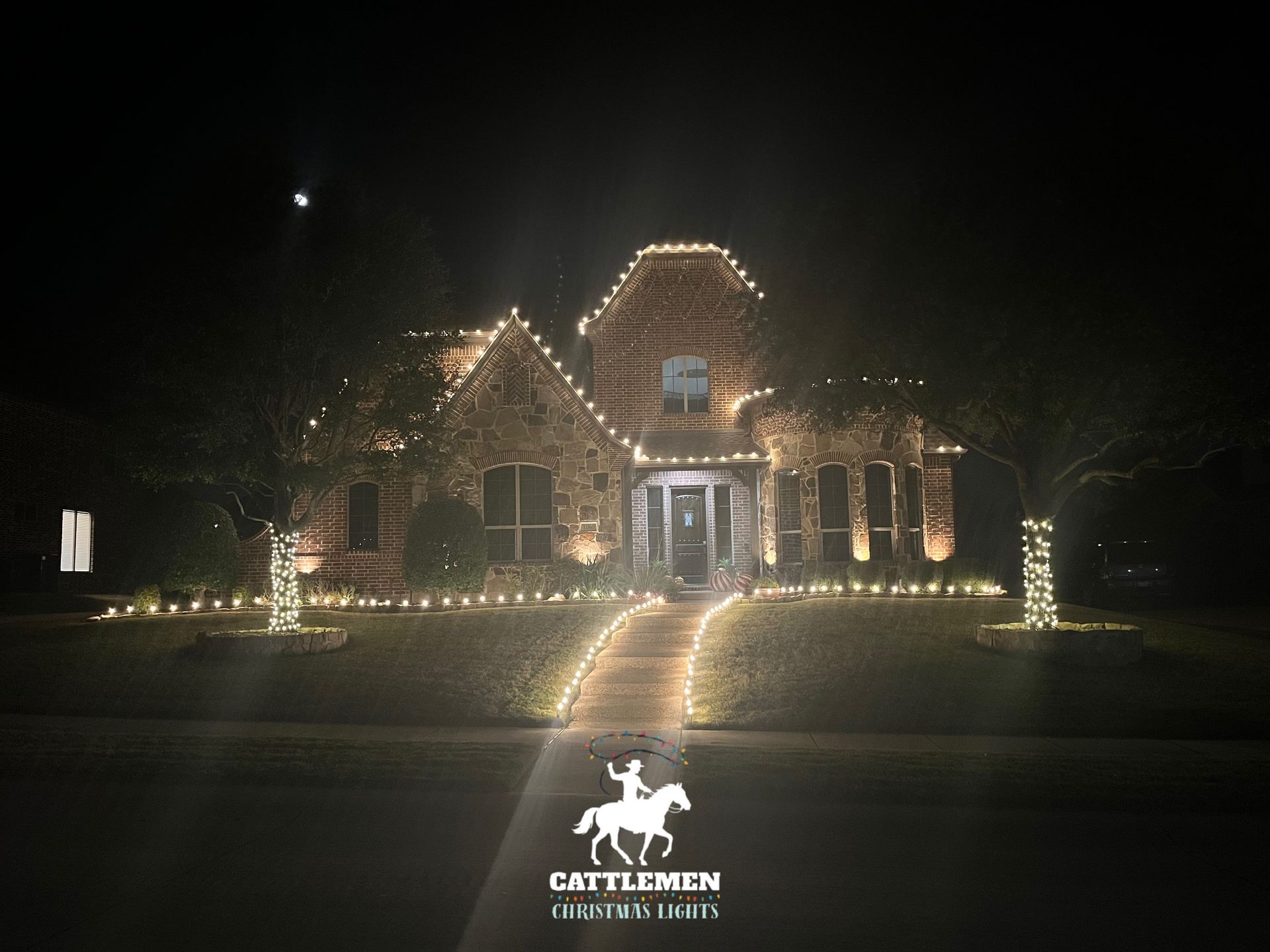 House outlined in white holiday lights at night with lit driveway and front yard decorations