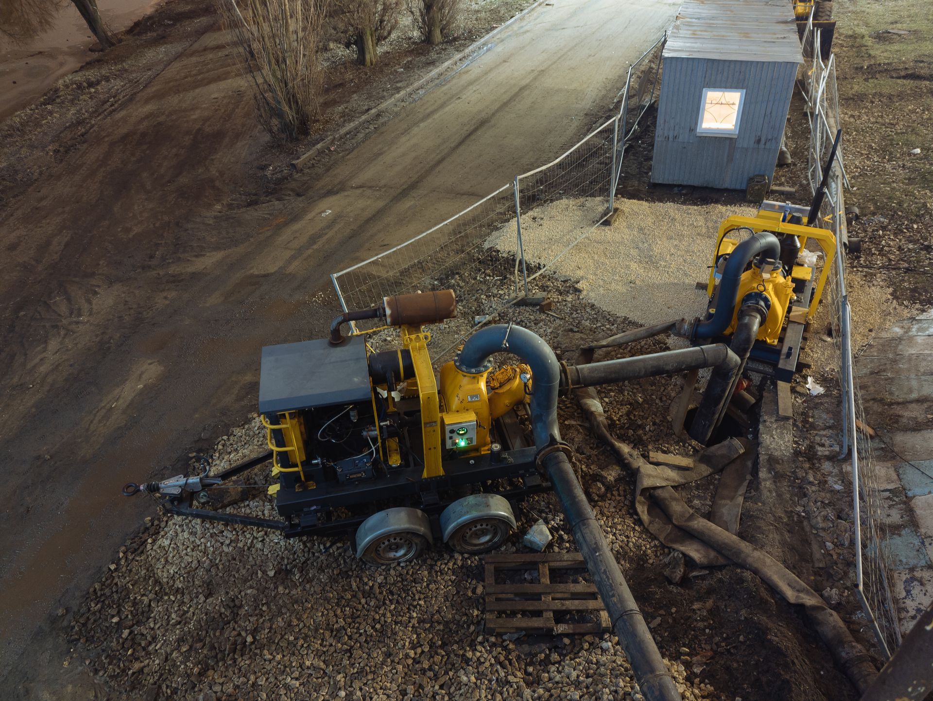 Two yellow industrial pumps on a gravel surface next to a road, with a small building in the background.