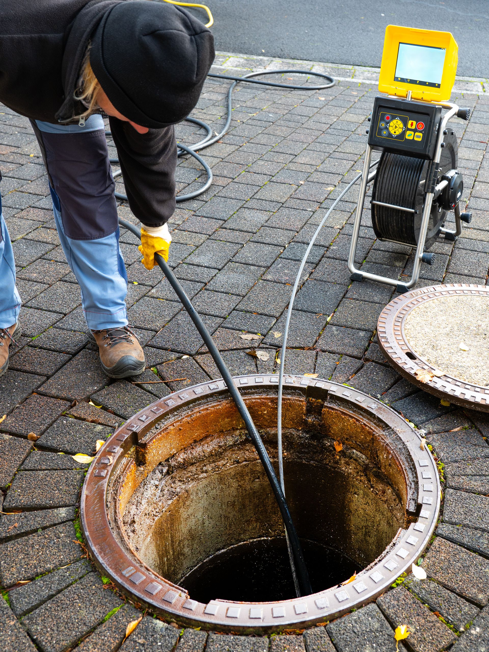 Person inspecting a sewer with a camera on a reel; open manhole in cobblestone street.