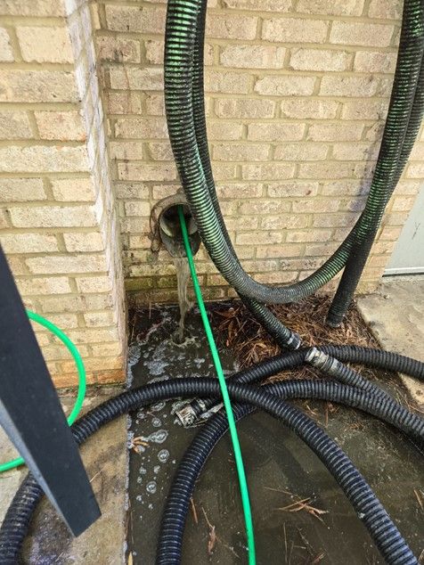 A green hose extends from a brick wall outlet, water flowing. Other hoses and debris are on the ground.