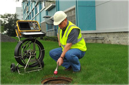Man in safety vest inspecting sewer with camera, equipment on grass.