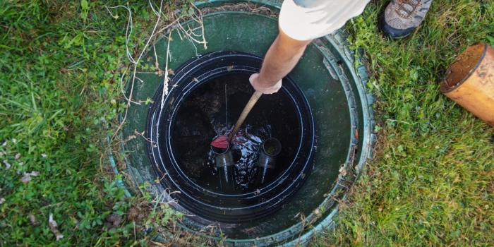 Person stirring contents in an open septic tank with a tool. Green grass surrounds the tank.