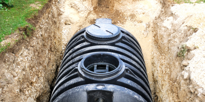 A black septic tank sits in a brown trench in the ground with a green grassy area beside it.