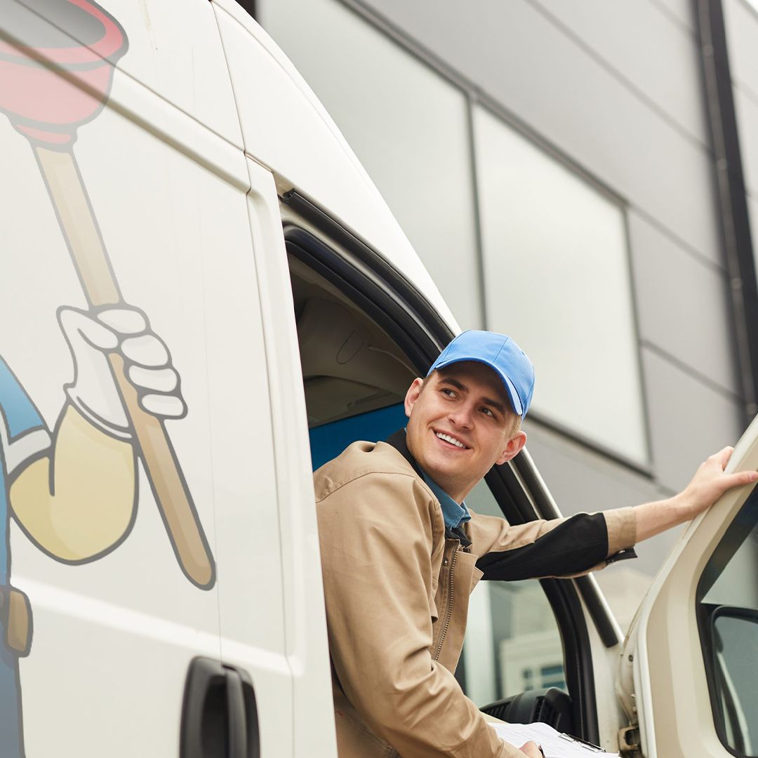 Plumber exiting a white van with a cartoon graphic, smiling, wearing a blue cap and tan jacket.