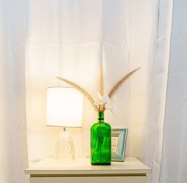 A green vase with flowers in it is on a table next to a lamp.