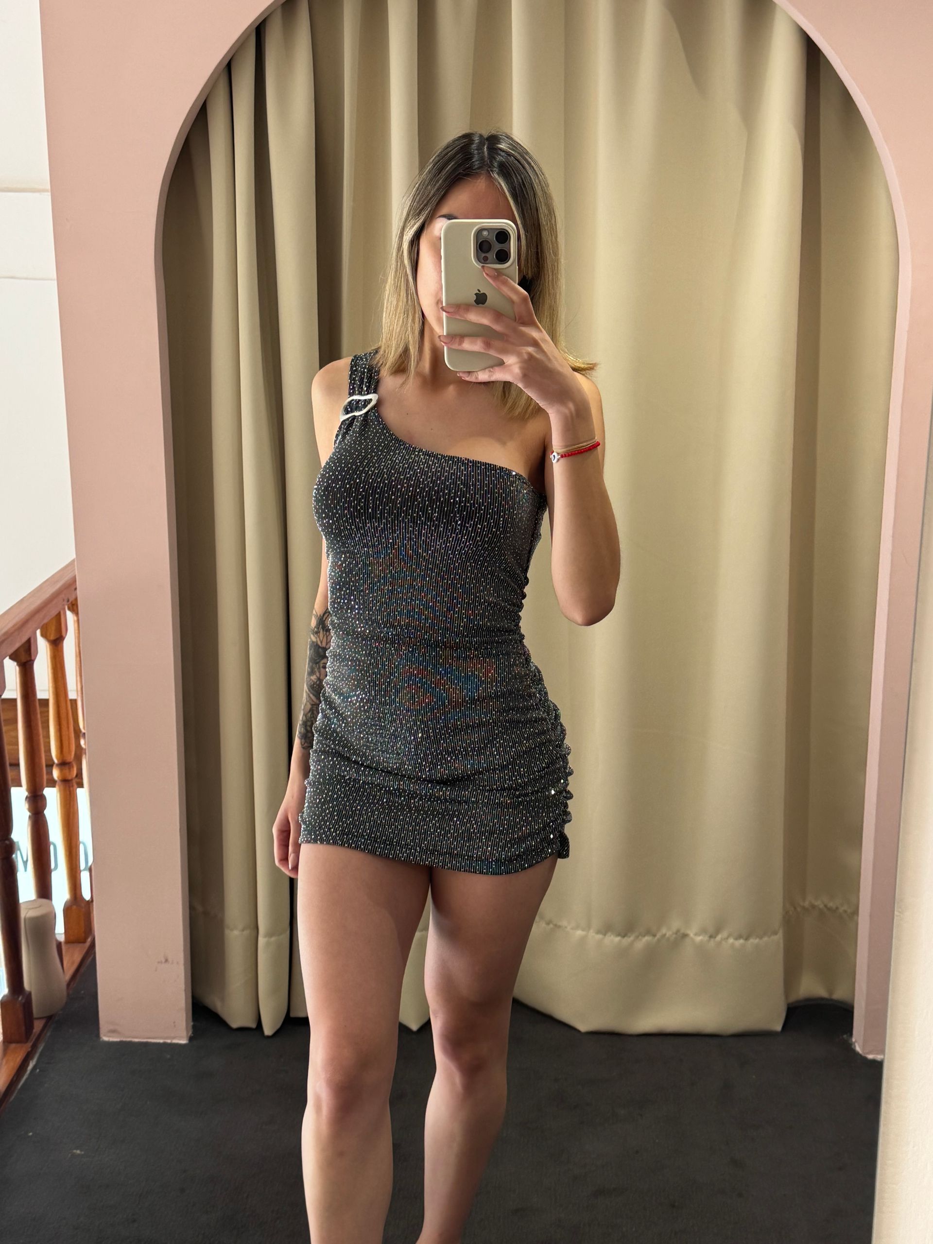Mujer con un minivestido brillante de un solo hombro tomándose una selfie frente al espejo en un probador.