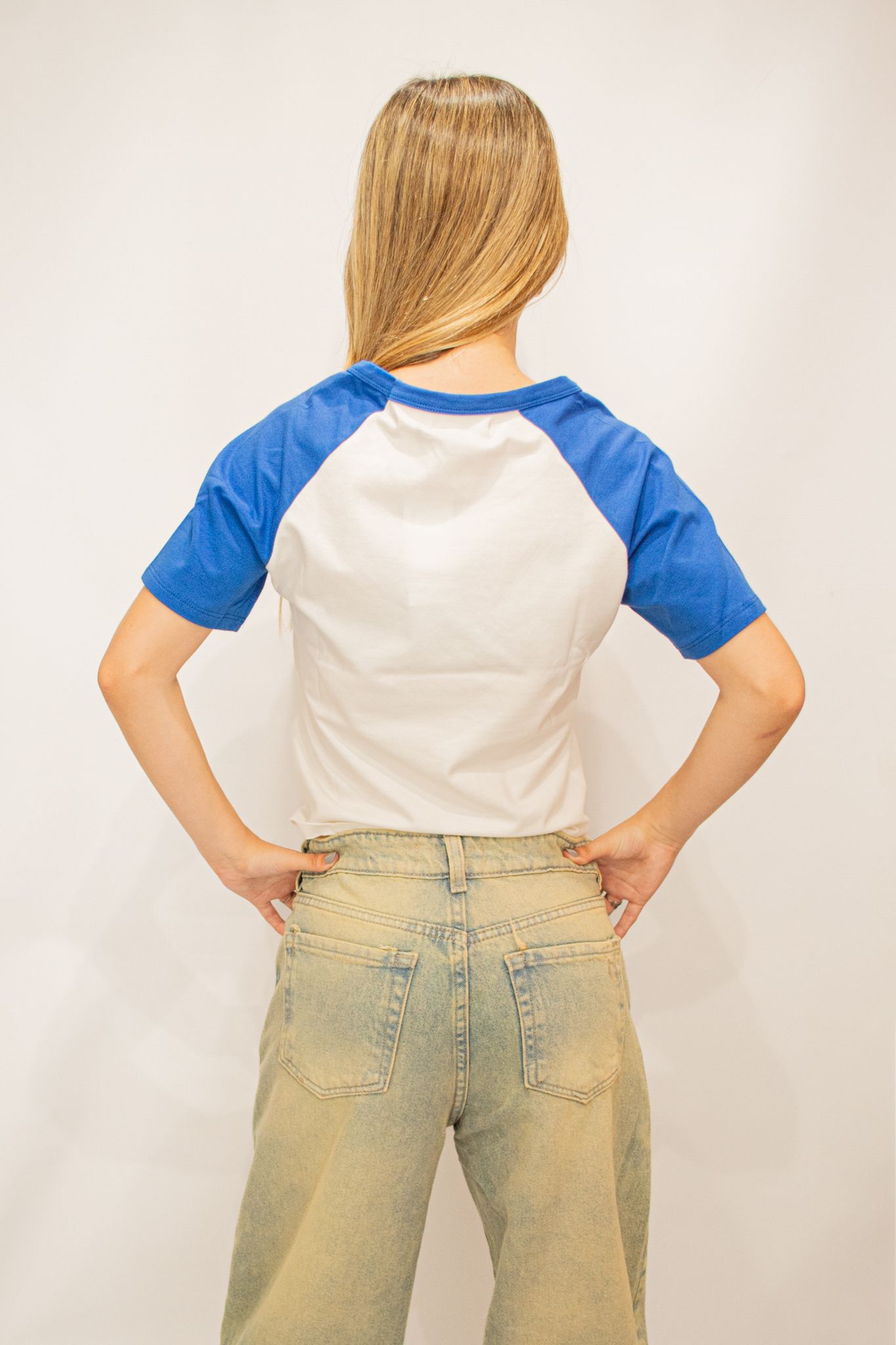 Mujer con camiseta de béisbol azul y blanca y pantalones cortos de mezclilla, de pie frente a un fondo blanco.
