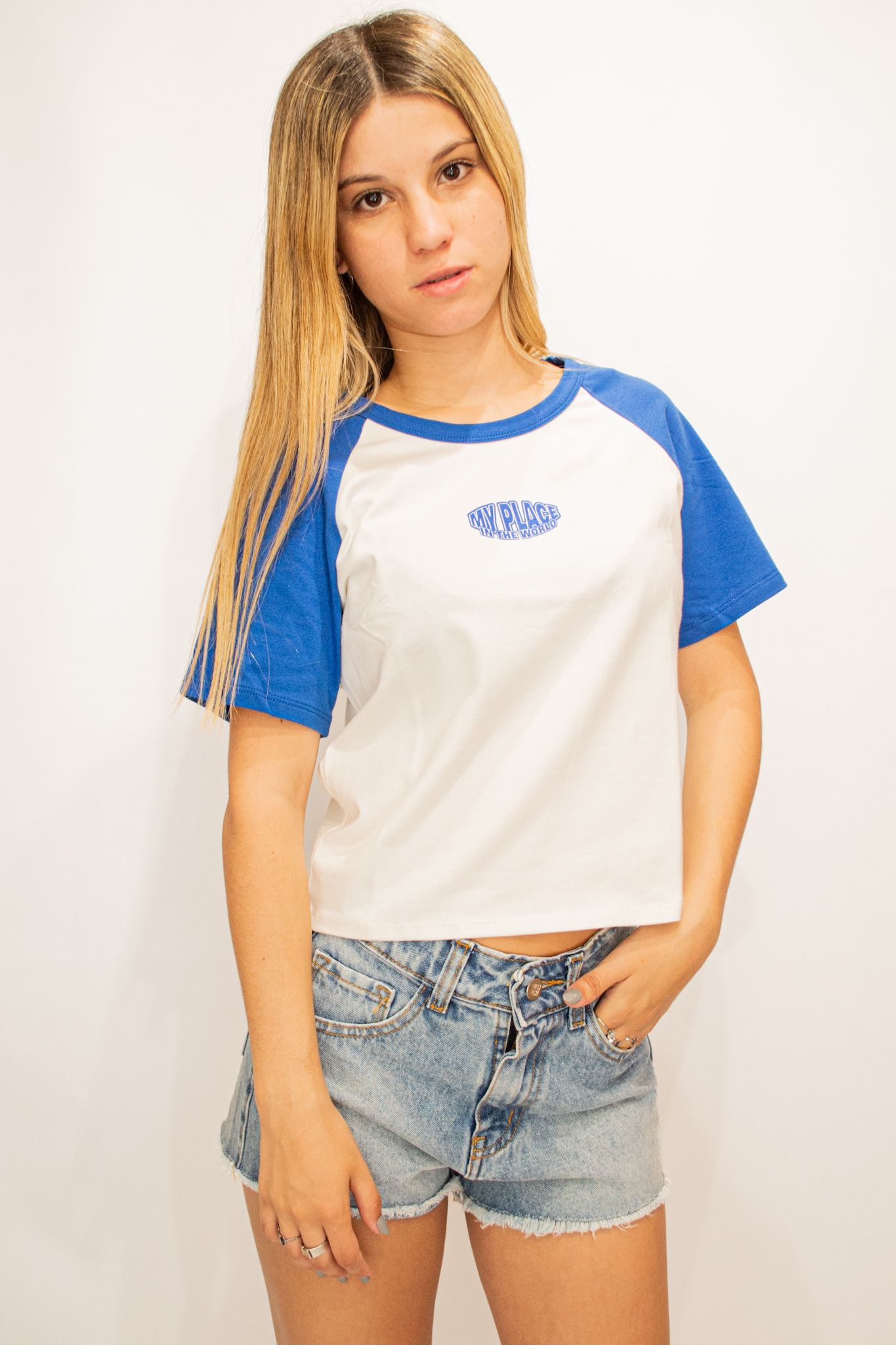 Mujer con camiseta de béisbol azul y blanca y pantalones cortos de mezclilla, de pie frente a un fondo blanco.