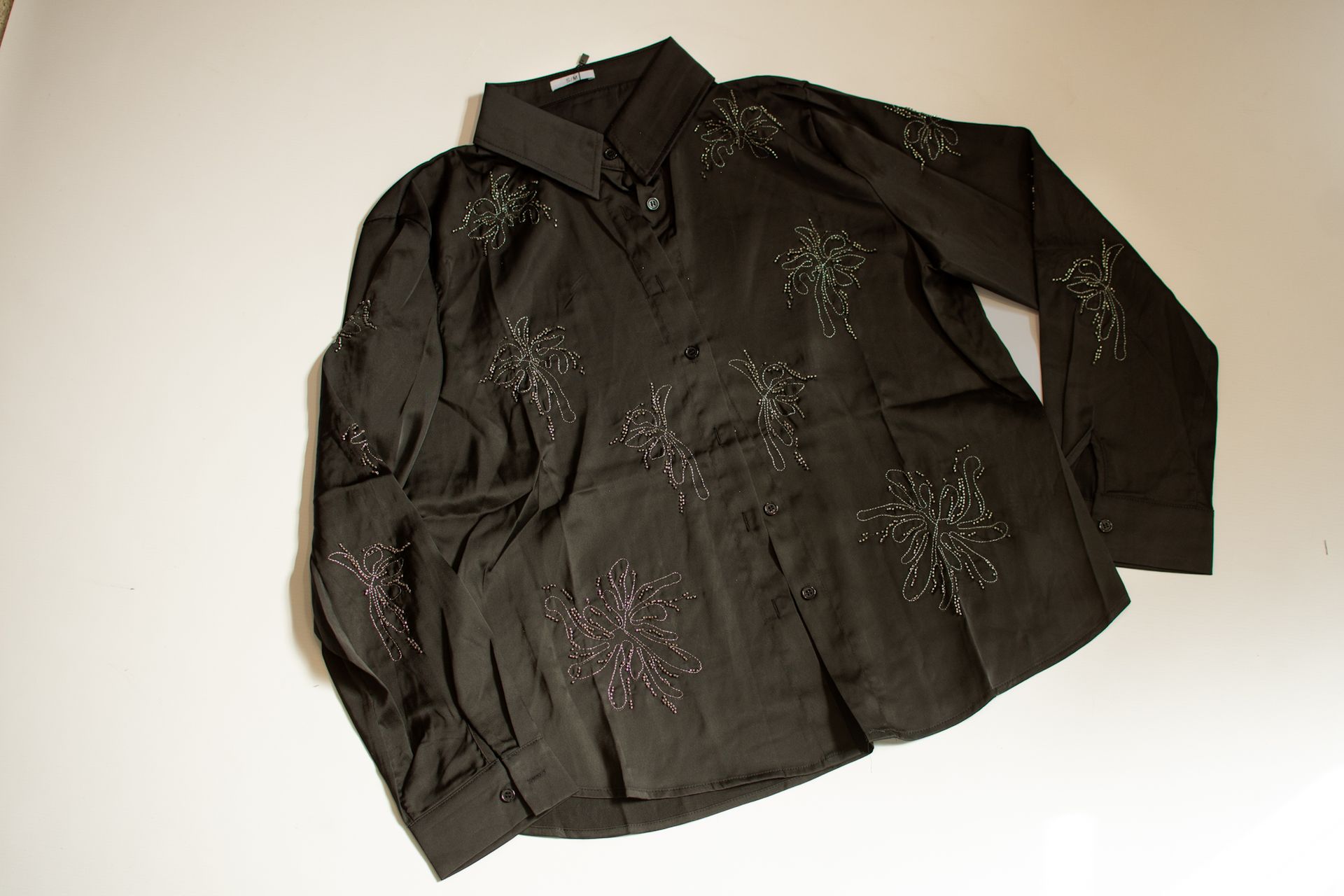 Camisa negra abotonada con adornos brillantes, colocada sobre una superficie blanca.