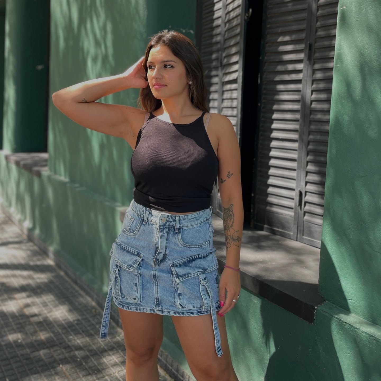Mujer con top negro y falda vaquera posa frente a una pared verde.