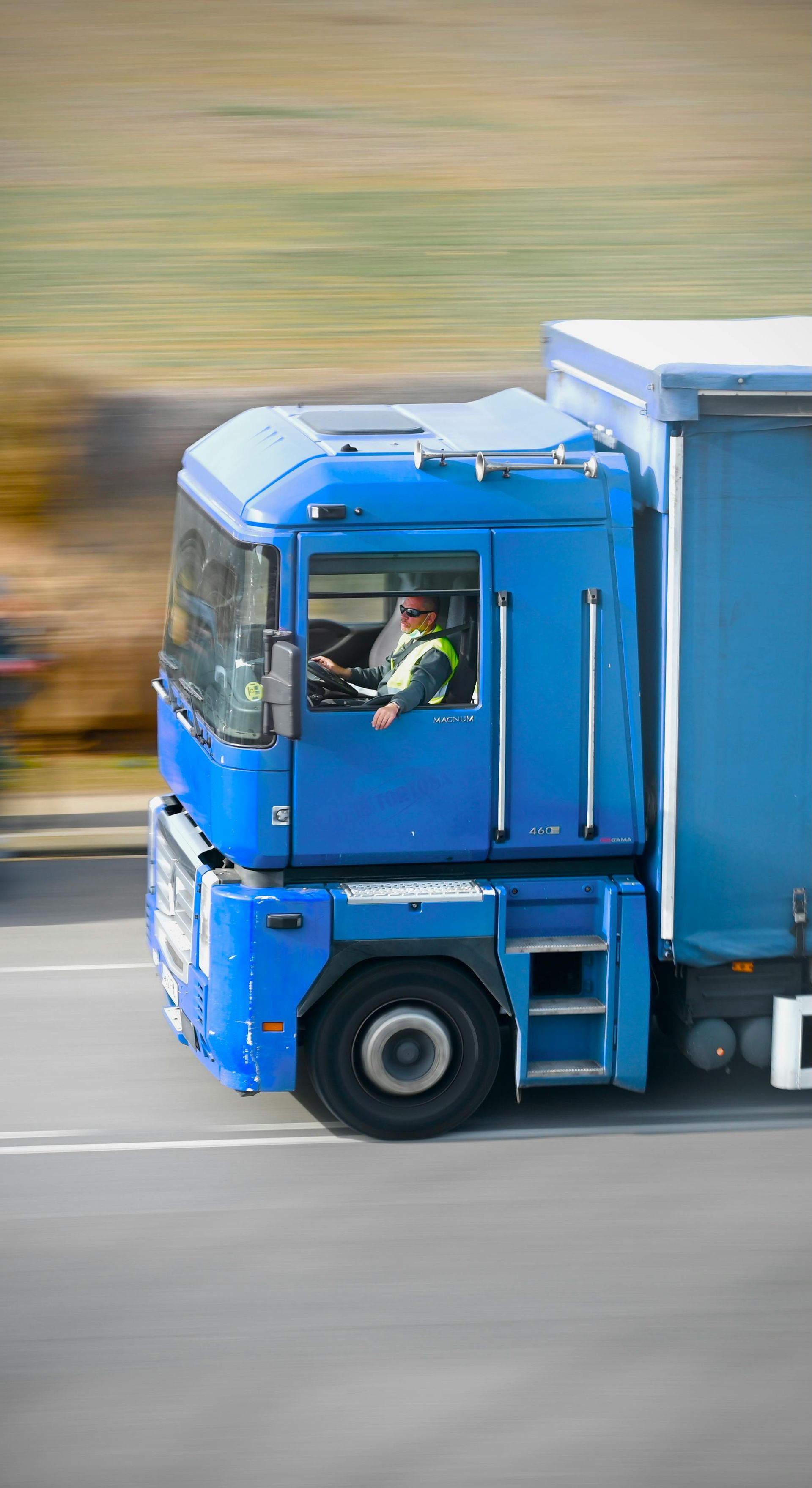 Blue semi-truck driving on a road, blurred background, driver visible in cab