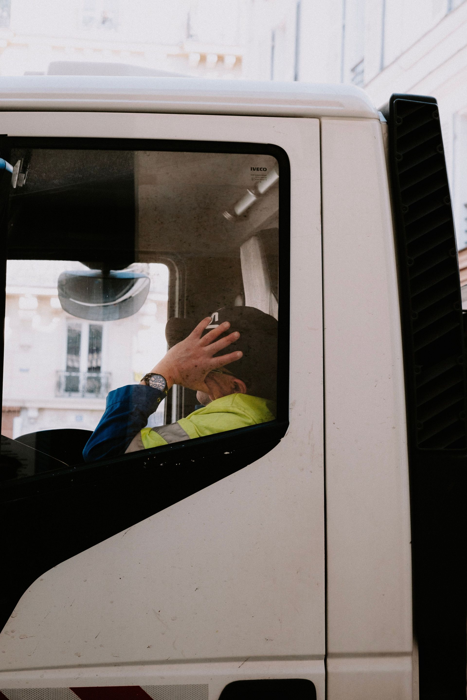 Person in a white truck cab, hand raised near the window, wearing a yellow safety vest.