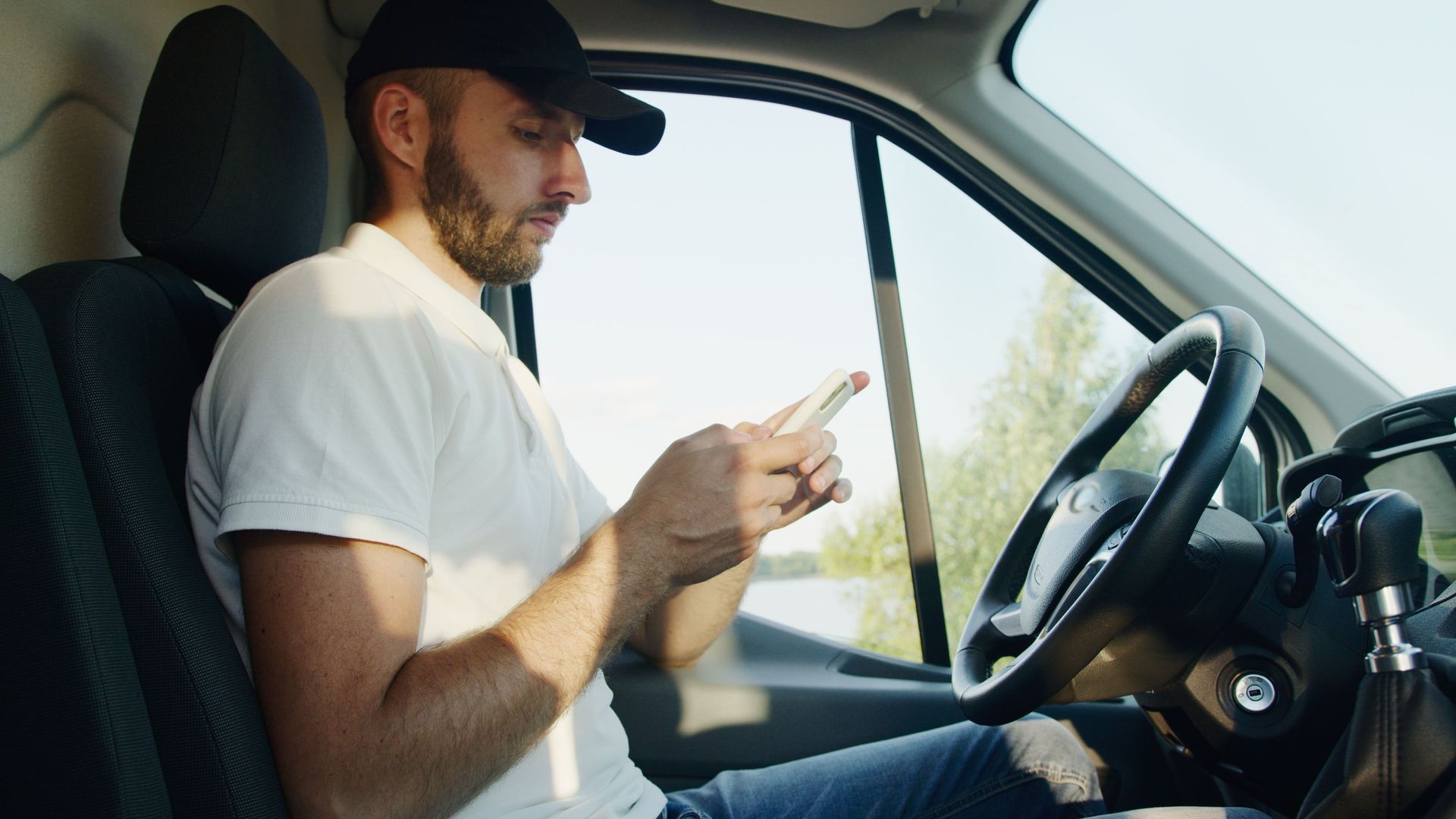 Man in a white T-shirt and black cap using a phone while sitting in a car driver’s seat