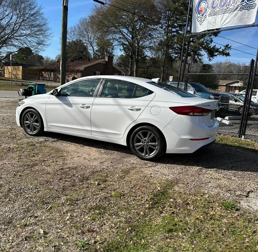 White sedan parked on a gravel lot beside a fenced street under sunny trees