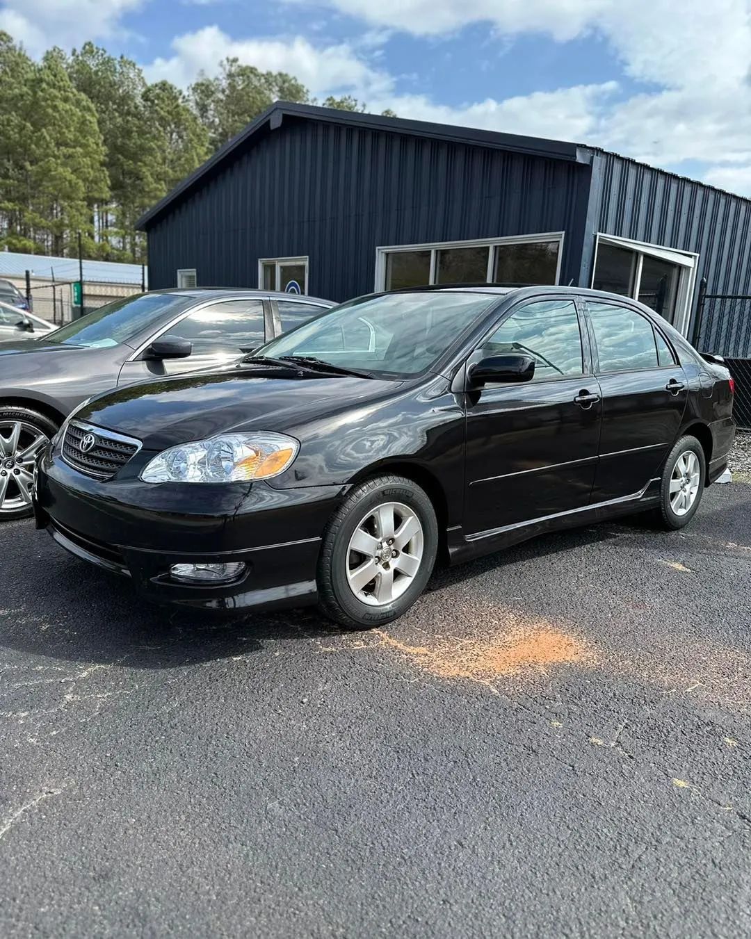 Black sedan parked beside a dark metal building on a gravel lot under a cloudy sky.