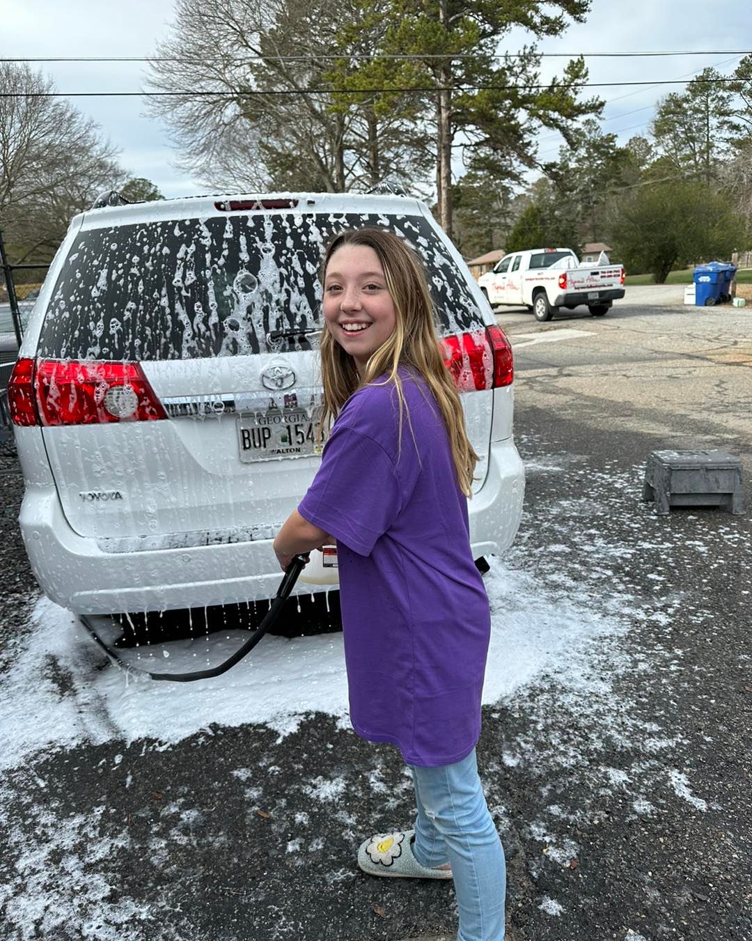 A young girl is washing a white suv with a high pressure washer.